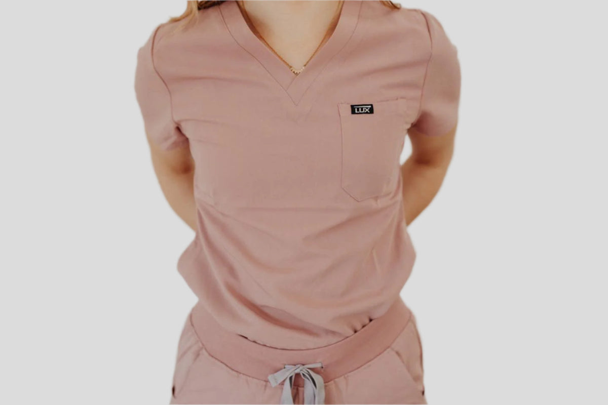 A woman wearing a pink V-neck scrub top with a logo on the chest pocket and a drawstring waist.