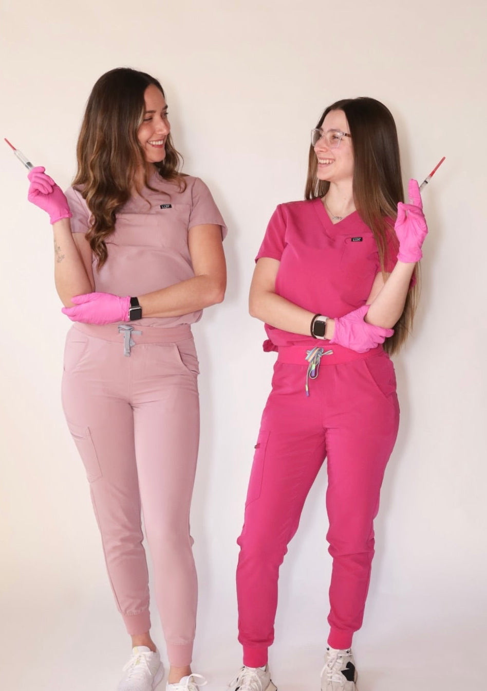 two women looking at eachother in lux scrubs jogger style scrub sets with syringes and pink gloves 